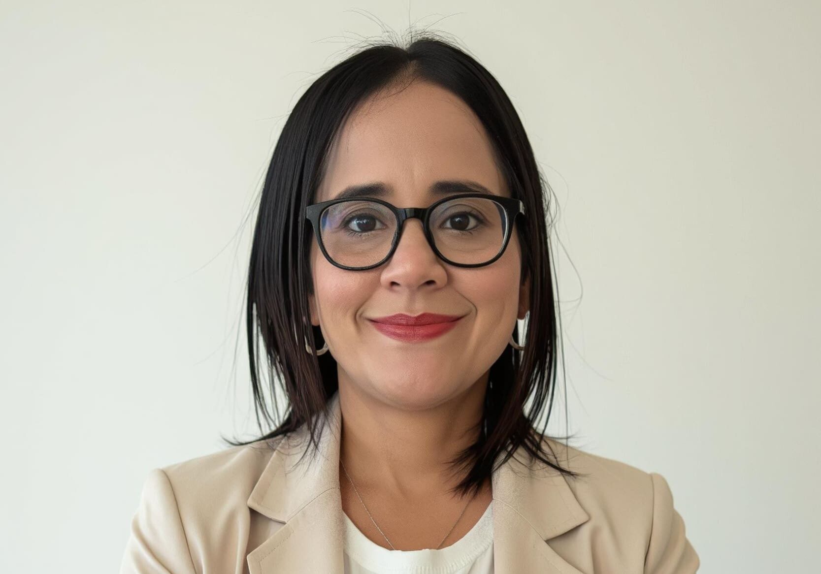 Smiling woman with short dark hair and black glasses, wearing a beige blazer, against a light background.