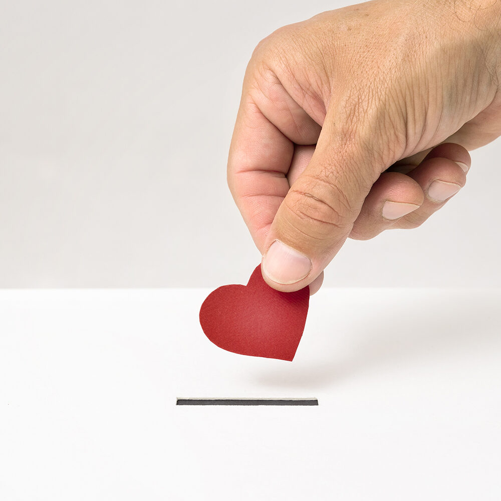 Red heart symbol is put by person’s hand into slot of white donation box