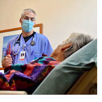 A doctor wearing a surgical mask chats with an elderly patient in a hospital bed