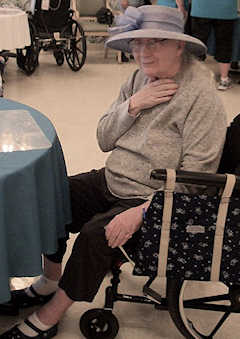 An elderly woman wearing a fancy hat is seated at a table in a care facility